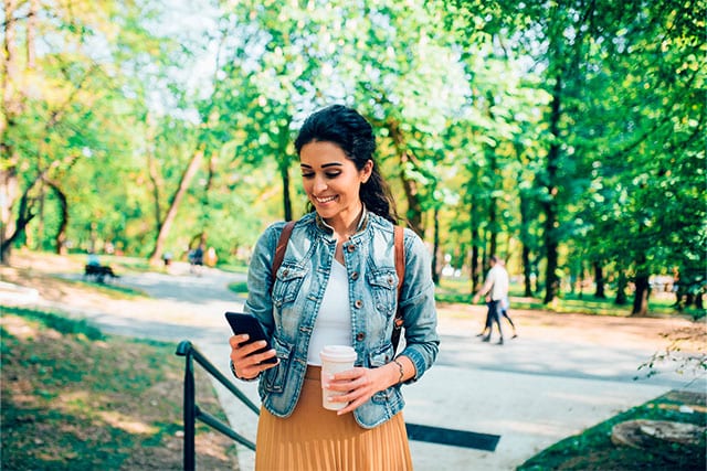 Una mujer caminando por un parque, sosteniendo una taza de café y revisando su teléfono celular.