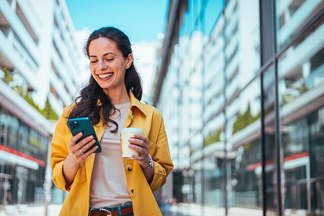 Una mujer caminando por la calle, sosteniendo un café y revisando su teléfono celular.