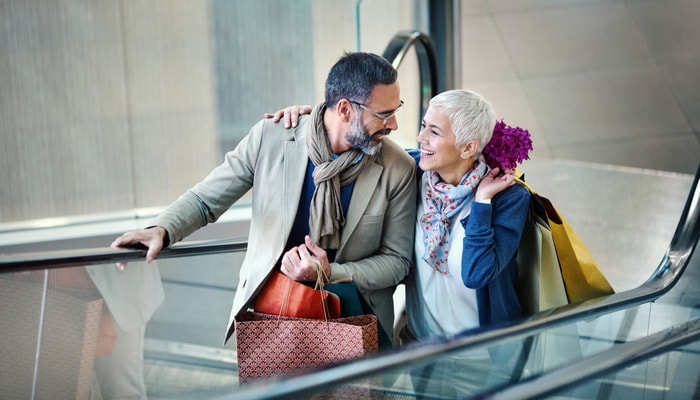 Pareja subiendo una escalera mecánica en un centro comercial. Llevan bolsas de compras y están contentos con la ropa que acaban de comprar.