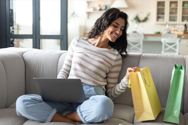 Mujer sentada en el sofá de su casa con una laptop en el regazo y dos bolsas con compras a su lado.