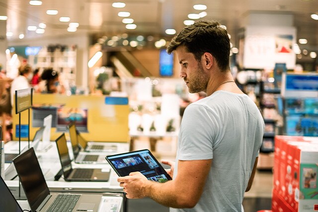 Hombre revisando laptops en una tienda departamental, sosteniendo una tableta.