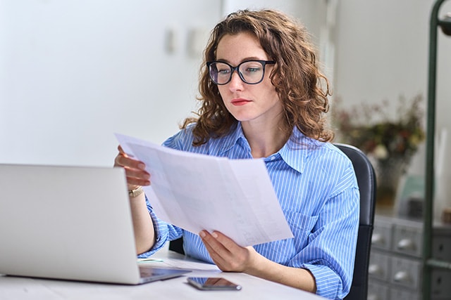 Mujer en casa revisando documentos junto a una laptop.