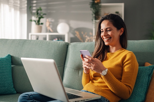 Mujer sentada en el sofá revisando su teléfono celular con una laptop en el regazo.