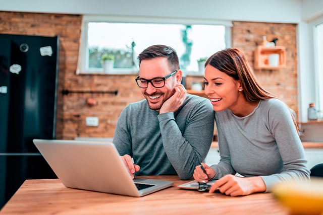 Una pareja sentada a la mesa de la cocina revisando una computadora laptop.