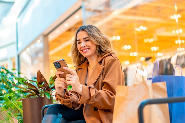 Una mujer revisa su teléfono sentada en un centro comercial cerca de sus bolsas de compras.