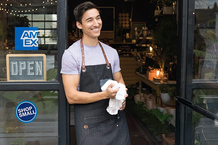 Dueño de un pequeño negocio en la puerta, con pegatinas visibles de Amex y Shop Small en el escaparate.