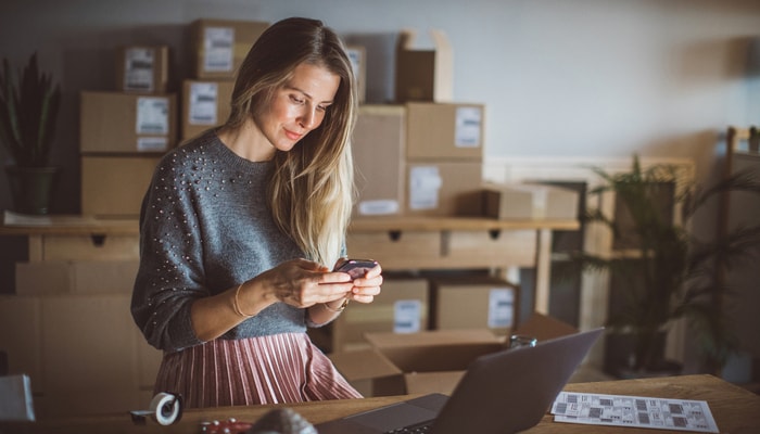 Mujer trabajando en una tienda online. Lleva ropa informal y usa su portátil y su celular para organizar su trabajo.