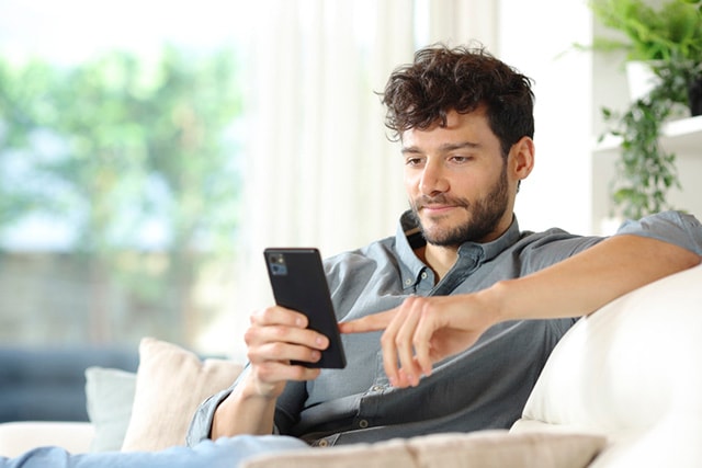 Un hombre sentado en el sofá de su casa mirando su celular.