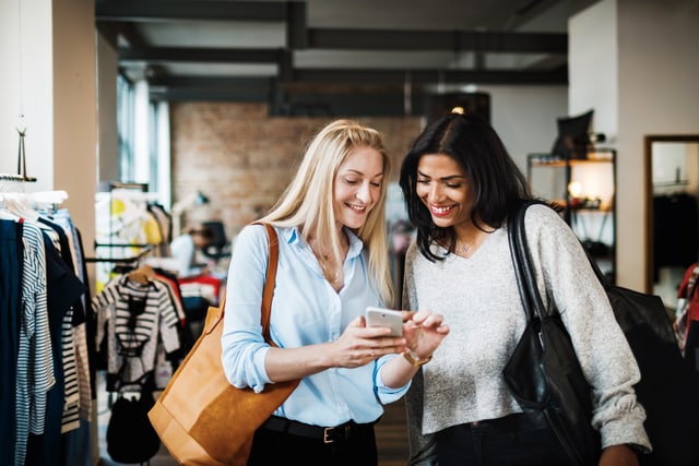 Dos mujeres sonriendo y mirando un celular mientras salen de compras juntas.