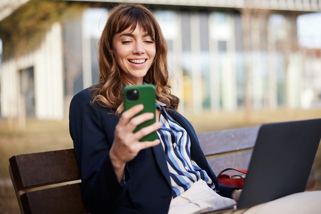Mujer sonriendo utiliza su celular sentada en un banco.