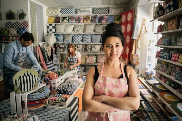 Mujer propietaria de una pequeña tienda textil posando dentro de su tienda con empleados al fondo.