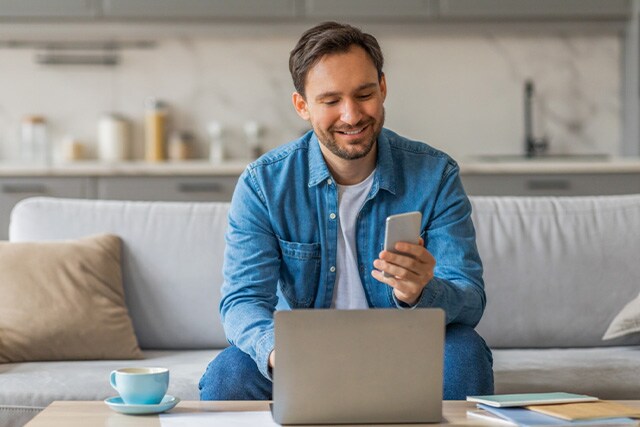 Hombre en casa, en el sofá, revisando una laptop mientras sostiene su teléfono celular.