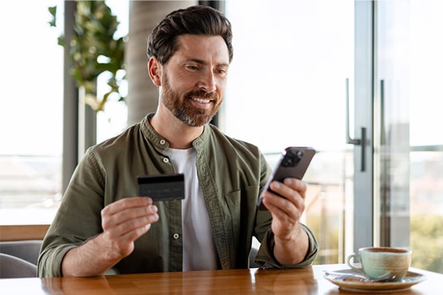 Hombre en una cafetería sosteniendo su teléfono celular y una tarjeta de crédito.