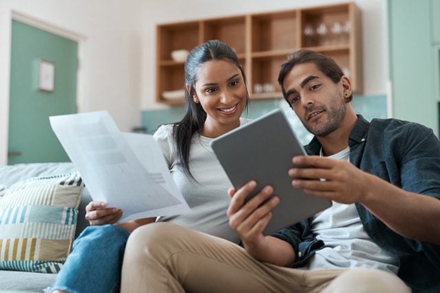 Una pareja en casa, sentada en el sofá, revisando papeleo juntos con una tablet.