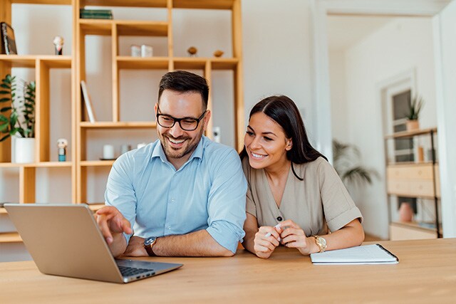 Una pareja en casa, sentada a una mesa y revisando sus finanzas en una computadora laptop.