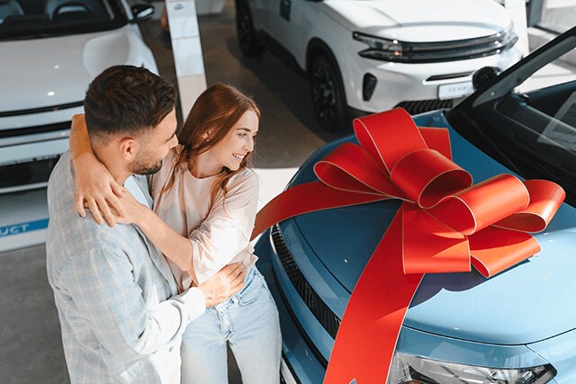 Pareja en una agencia de autos abrazándose junto a un auto nuevo con un lazo.