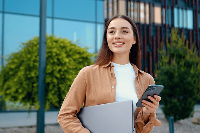 Mujer sosteniendo una laptop y su teléfono celular frente a un edificio de oficinas.