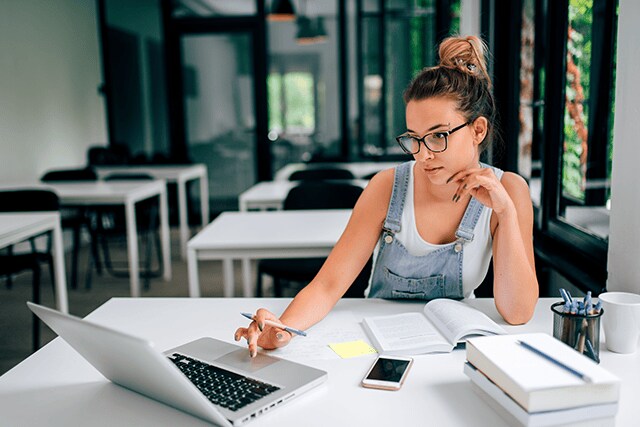 Mujer en una oficina trabajando en una laptop, con un cuaderno y algunos libros a su alrededor.