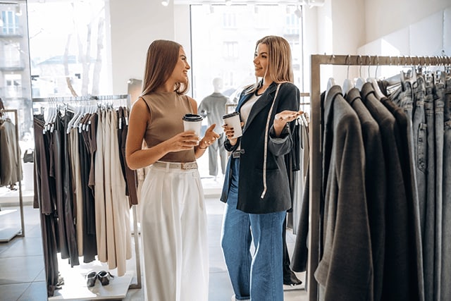 Dos amigas sosteniendo vasos de café en una tienda de ropa.