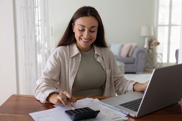 Mujer sonriente sentada en un escritorio con un ordenador portátil, documentos y una calculadora.