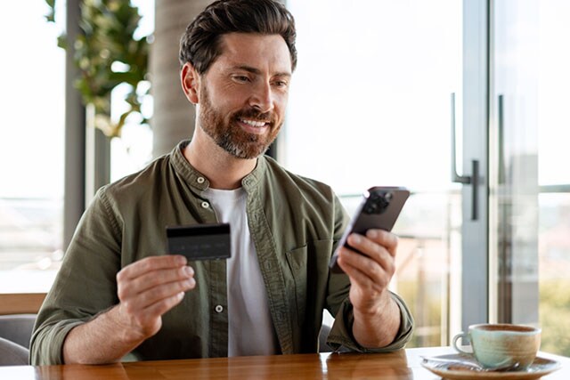 Un hombre en una cafetería sostiene su teléfono celular y una tarjeta de crédito.