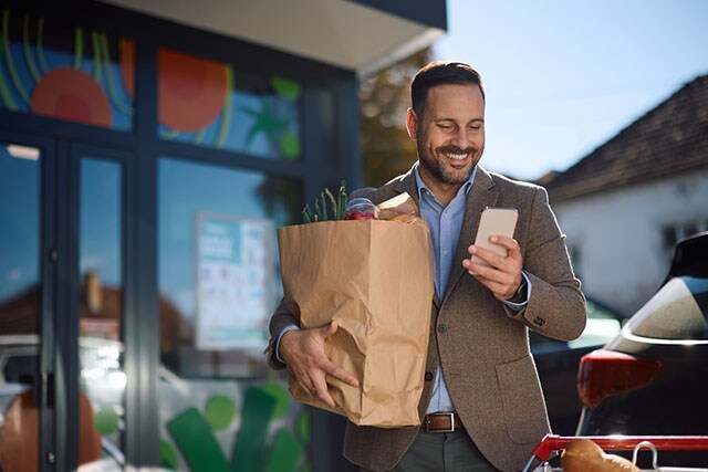 Un hombre con una bolsa de papel para la compra en la puerta de un supermercado, mirando su teléfono celular.