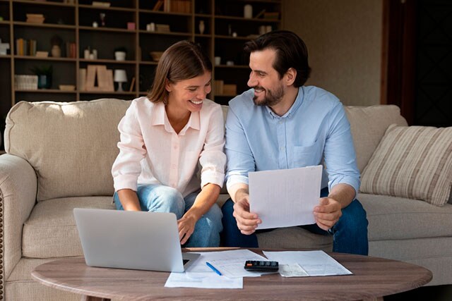 Una pareja sonriente sentada en el sofá revisando documentos con una computadora portátil.