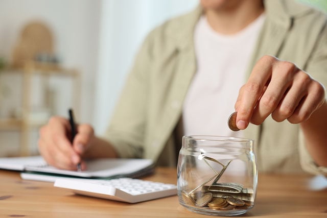 Una persona deposita monedas y cambio en un pequeño frasco, cerca de una calculadora y un bloc de notas.