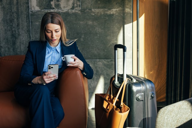 Mujer de negocios tomando café y usando un teléfono inteligente mientras espera con su equipaje en la sala VIP del aeropuerto.