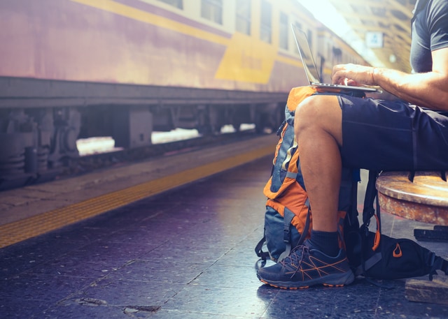 Mochilero nómada digital trabajando en la computadora mientras espera el tren.