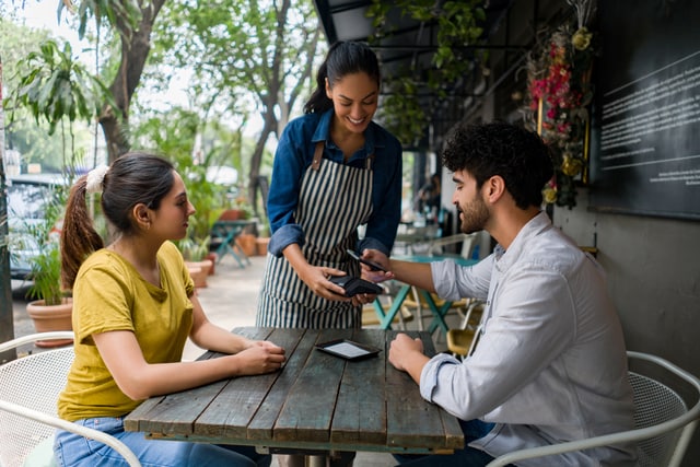 Hombre realiza un pago contactless con su teléfono celular a una camarera en un café.