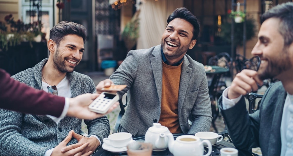 Men sitting around cups of coffee at outdoor dining, and one man paying with credit card