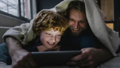 a man and a boy are laying under a blanket looking at a tablet .