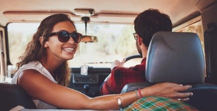 a man and a woman are sitting in a car. woman is turned back wearing sunglasses and smiling.