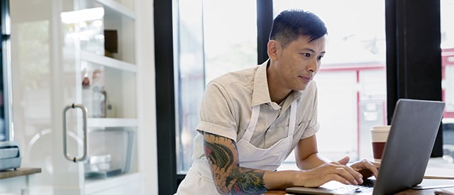 Une personne travaillant de la maison, assise à un bureau entourée de plantes. Un rayon de soleil perce derrière elle. Elle est au téléphone et regarde son ordinateur portable.