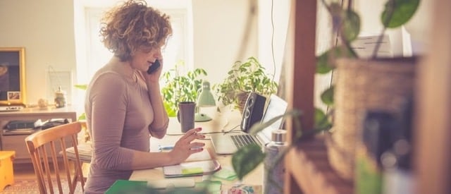 Une personne travaillant de chez elle, assise à un bureau entouré de plantes. Un rayon de soleil brille par la fenêtre derrière elle. Elle est au téléphone et consulte son ordinateur portable.