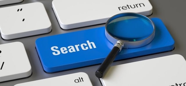 Une gros plan sur une touche de clavier d'ordinateur. Celle-ci est bleu vif entourée de touches blanches. Y figure le mot "search" (recherche) et y est posée une loupe miniaturisée.