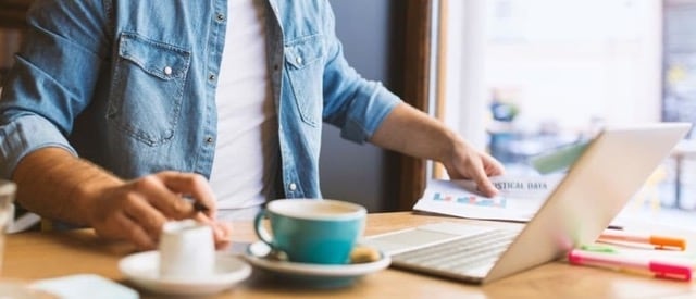 Personne travaillant d'un café. La journée est ensoleillée, une tasse et un laptop son posés sur la table devant elle.