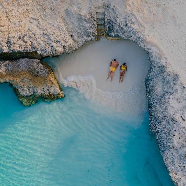 2 mensen liggen op een strand in een baai omgeven door rotsen en een azuurblauwe zee