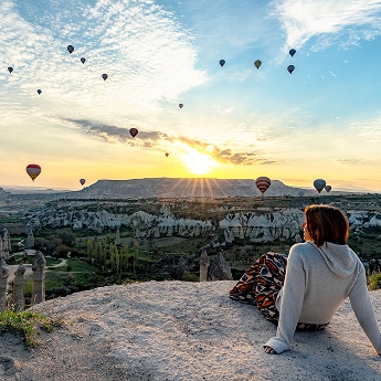 Vrouw op een rotsformatie kijkt uit over een vallei in Cappadocië vol heteluchtballonnen bij zonsopgang.
