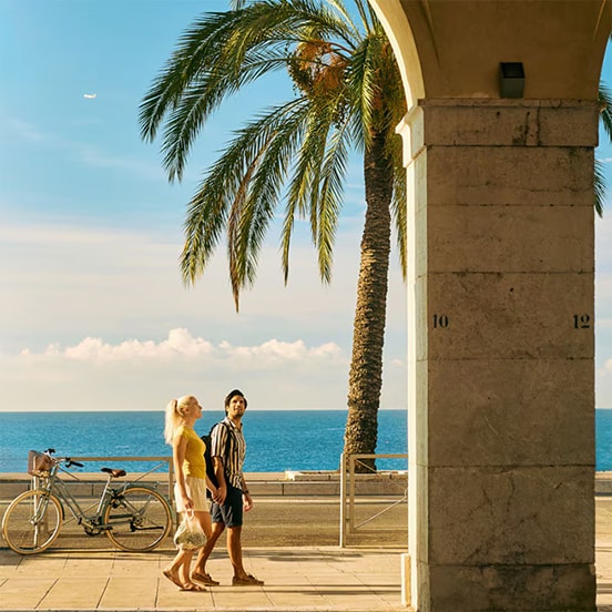 Man en vrouw lopen hand in hand langs de zee onder een grote palmboom en een klassieke stenen arcade.