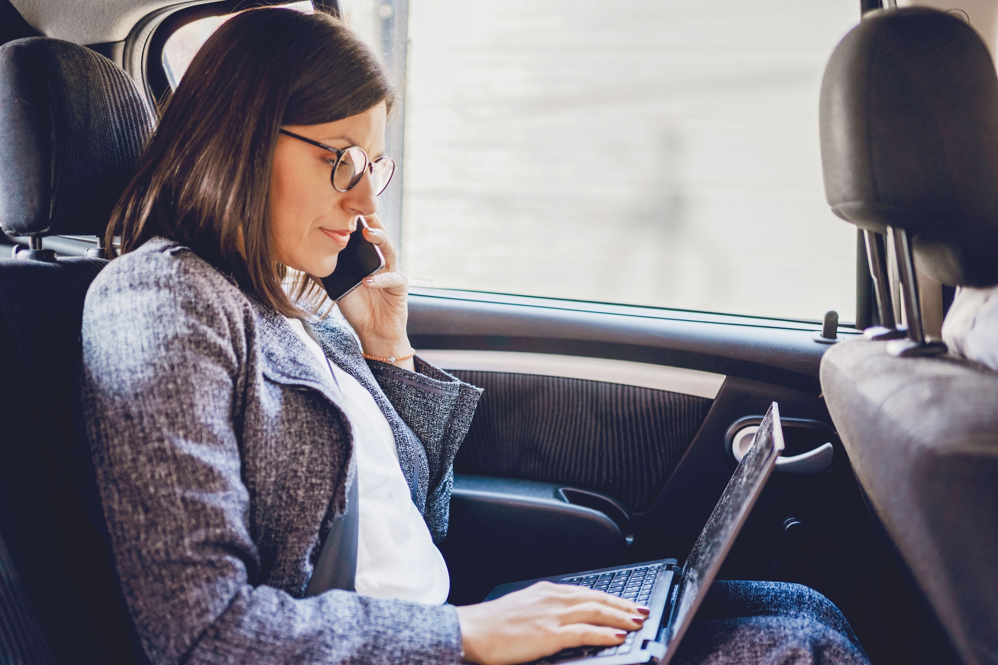 Business woman working in a car. She is talking on the phone and using a laptop.