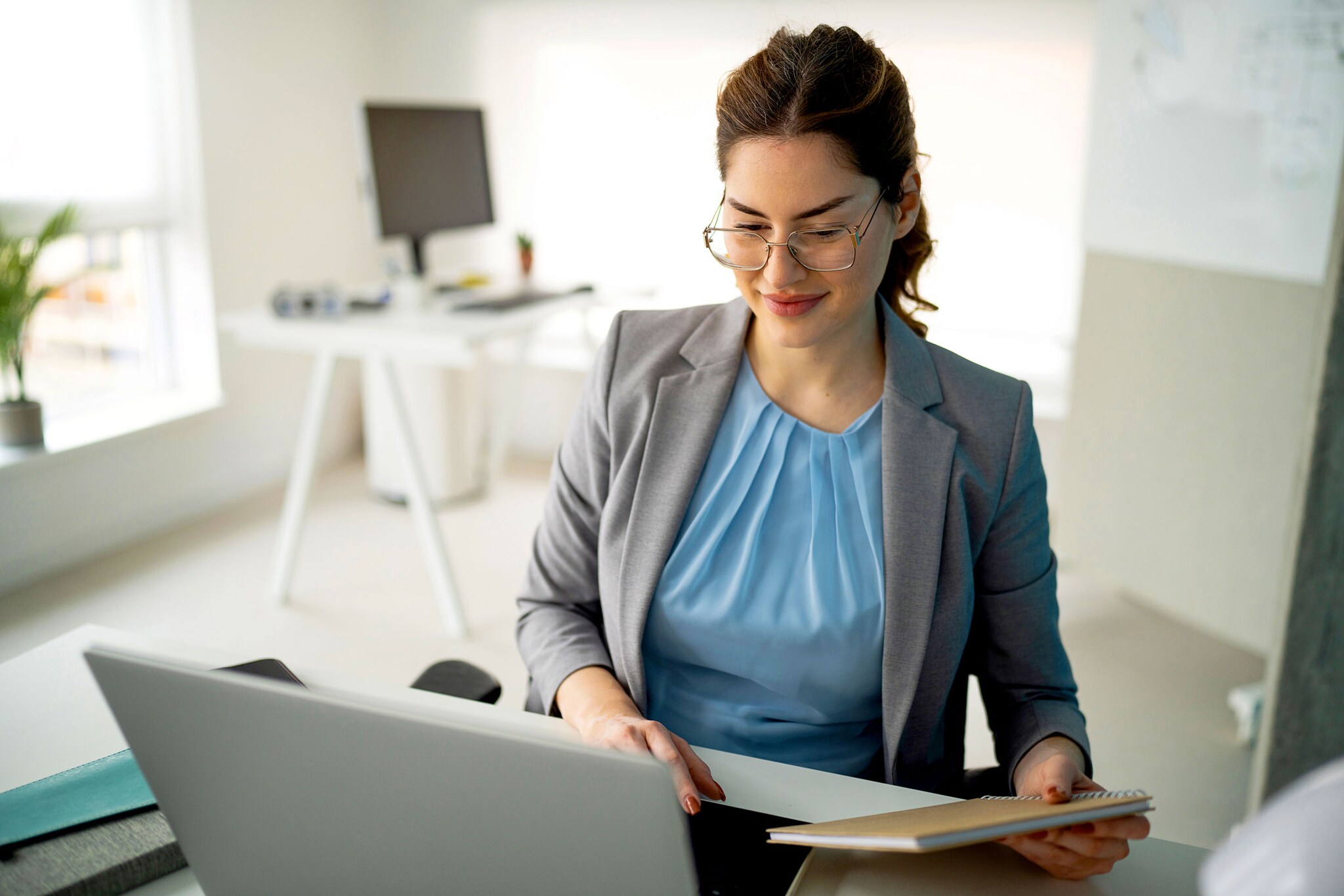 A young woman is seated at a desk in a modern office environment, focused on her laptop while holding a notebook. She wears glasses and a professional outfit, with a bright and airy workspace in the background.