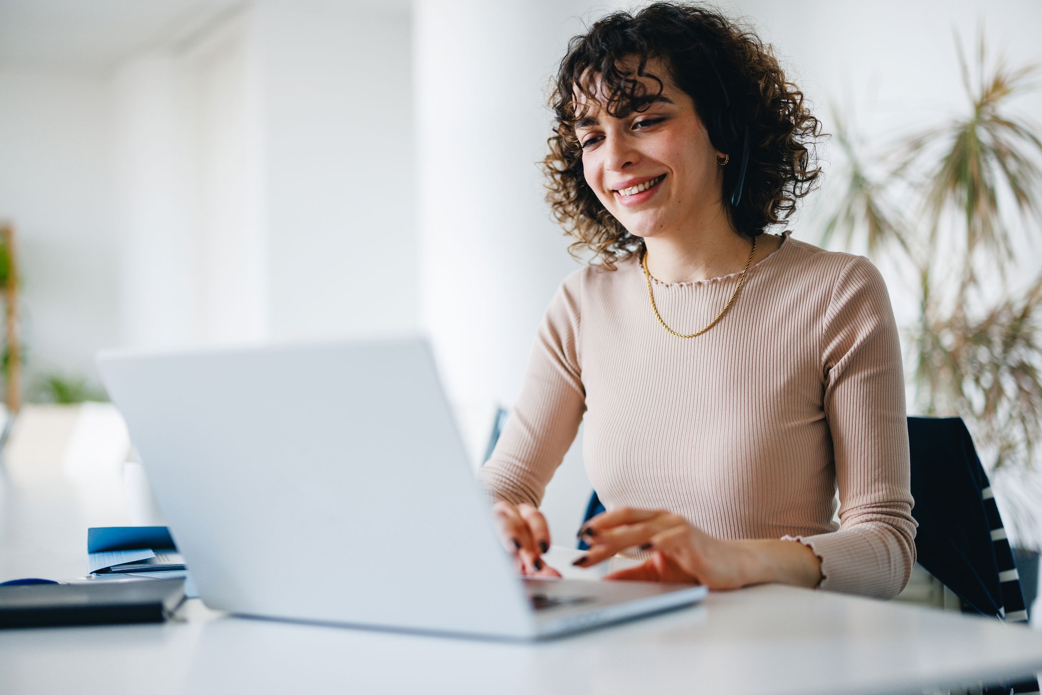 Young adult woman smiling while working on her laptop at a bright modern office desk.