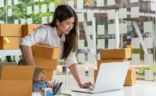 Woman standing over desk on laptop holding boxes