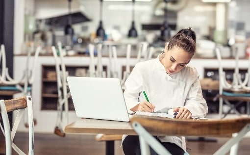 Chef of a restaurant sitting at a table with her laptop and writing in a book
