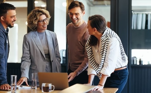 Four coworkers standing and having a discussion near a table with a laptop
