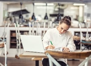 Lady sitting at table in an empty restaurant with laptop and notebook