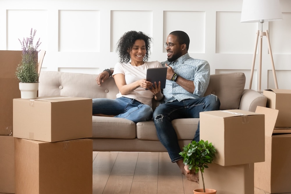 two people sitting on couch looking at tablet surrounded by moving boxes
