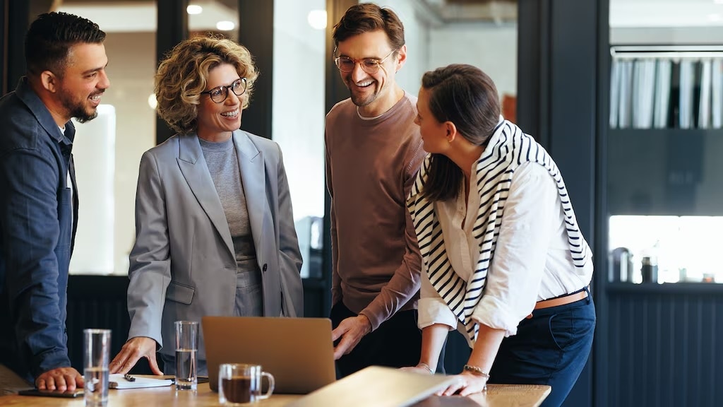 Four individuals standing around a table looking at laptop